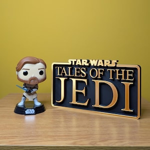 May include: A Star Wars collectible featuring an Obi-Wan Kenobi figure with a lightsaber and a sign that reads "Star Wars Tales of the Jedi". The sign has gold lettering on a black background.