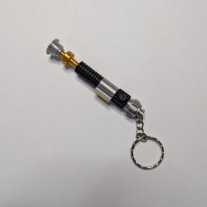 May include: A miniature lightsaber keychain with a silver, black, and gold design. The keychain has a ring attached for keys. The lightsaber has a detailed handle and a cylindrical shape.