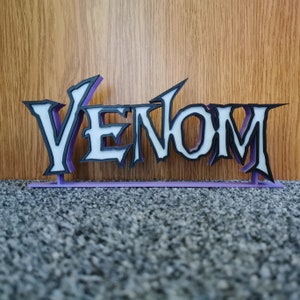 May include: A 3D-printed sign displaying the word "VENOM" in a distinctive font. The letters are white with black edging, set against a purple backing and base. The sign is placed in front of a wooden panel.