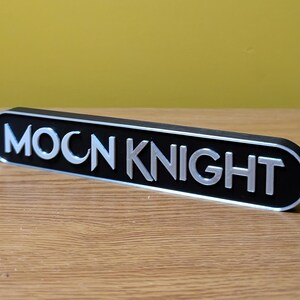 Puede incluir: Un letrero de metal negro y plateado con las palabras "MOON KNIGHT" en letras plateadas.
