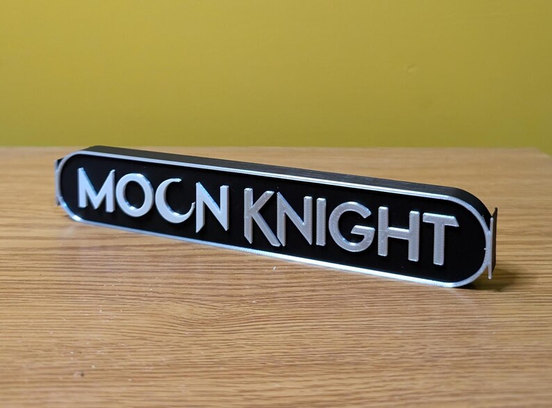 Puede incluir: Un letrero de metal negro y plateado con las palabras "MOON KNIGHT" en letras plateadas.