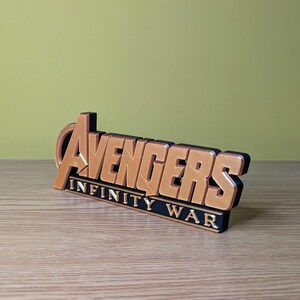 May include: A 3D-printed Avengers Infinity War logo in gold and black. The logo is angled slightly, with the words "Avengers" in a bold, stylized font above "Infinity War". The item is displayed on a wooden surface.