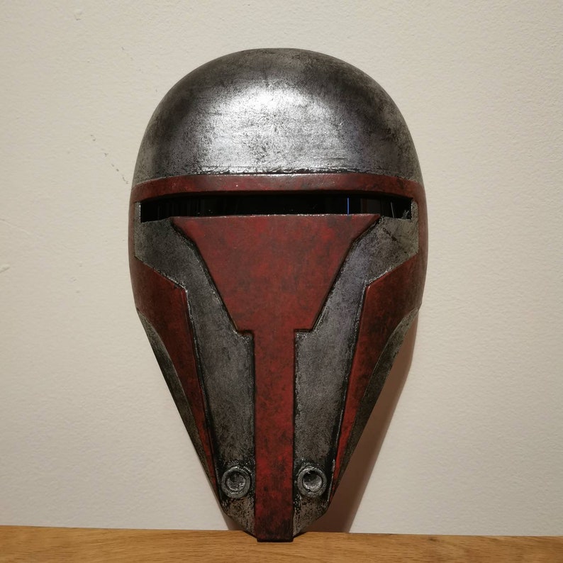May include: A red and silver Mandalorian helmet with a weathered finish. The helmet has a visor and two circular details on the bottom.
