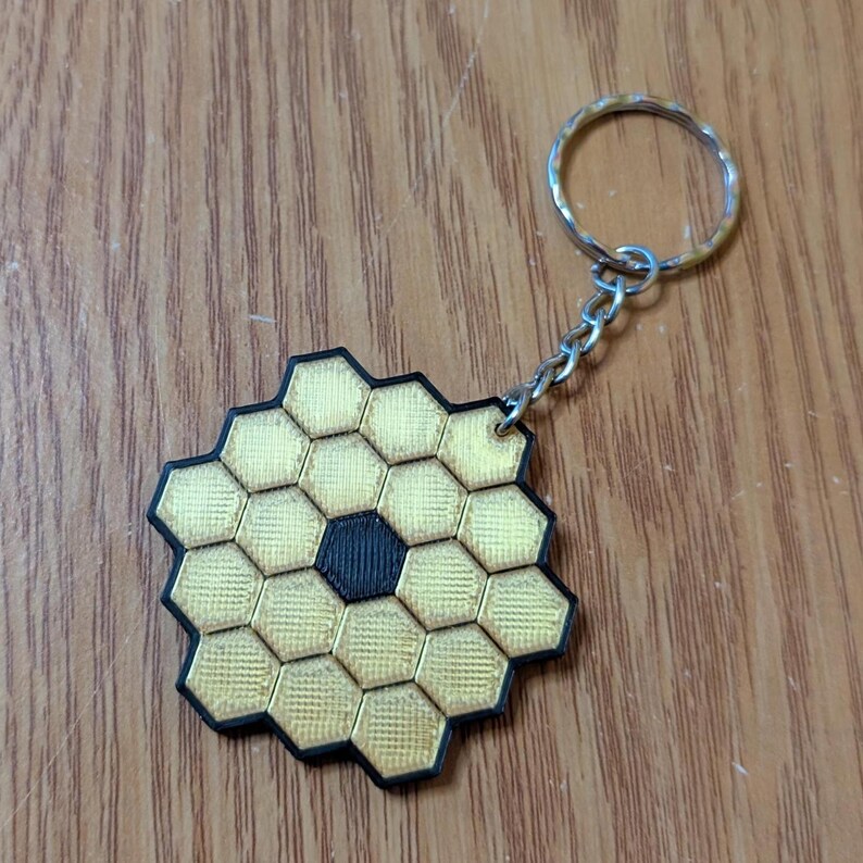 May include: A gold and black keychain featuring a honeycomb design with a central black hexagon. The keychain has a silver chain and ring. The design is reminiscent of the James Webb Space Telescope.