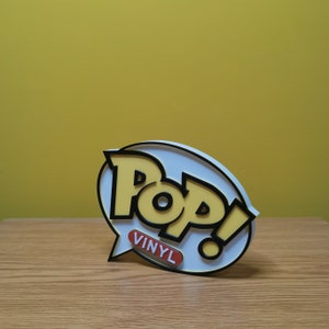 May include: A white and yellow 3D Pop! Vinyl logo with black outlines. The logo is on a white background and has a speech bubble with the word "Vinyl" in orange.