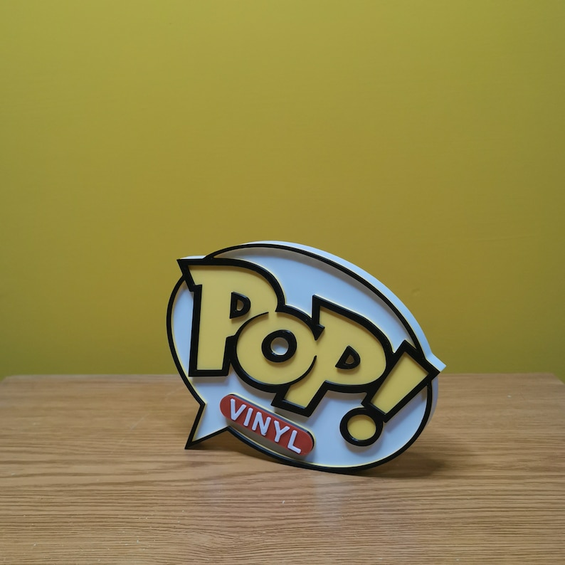 May include: A white and yellow 3D Pop! Vinyl logo with black outlines. The logo is on a white background and has a speech bubble with the word "Vinyl" in orange.