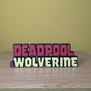 Może przedstawiać: Tr&oacute;jwymiarowy znak z napisem "DEADPOOL & WOLVERINE" w czerwonych i ż&oacute;łtych literach.