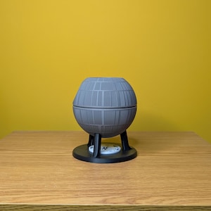 May include: A gray, spherical Death Star-shaped planter with a black base and three legs. The planter is made of a textured material and has a detailed design.