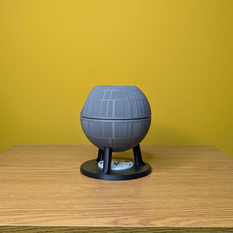 May include: A gray, spherical Death Star-shaped planter with a black base and three legs. The planter is made of a textured material and has a detailed design.