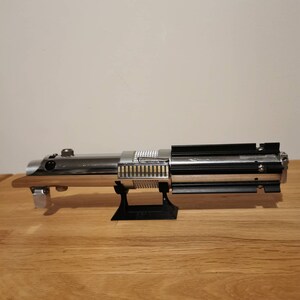 May include: A detailed replica lightsaber, featuring a polished chrome finish with black and silver accents. The hilt has a wooden grip and is displayed on a black stand. The lightsaber is a prop from the Star Wars franchise.