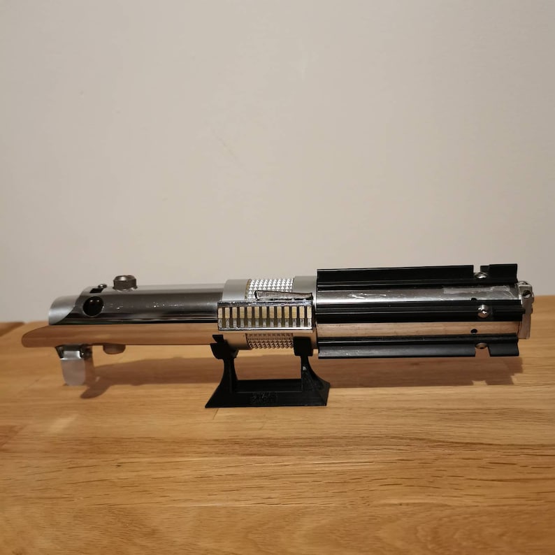 May include: A detailed replica lightsaber, featuring a polished chrome finish with black and silver accents. The hilt has a wooden grip and is displayed on a black stand. The lightsaber is a prop from the Star Wars franchise.