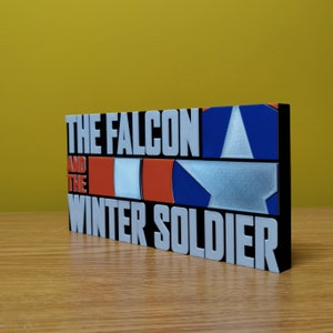 May include: A black and white 3D printed sign with the text "The Falcon and the Winter Soldier" in white letters. The sign features a red, white, and blue American flag design.