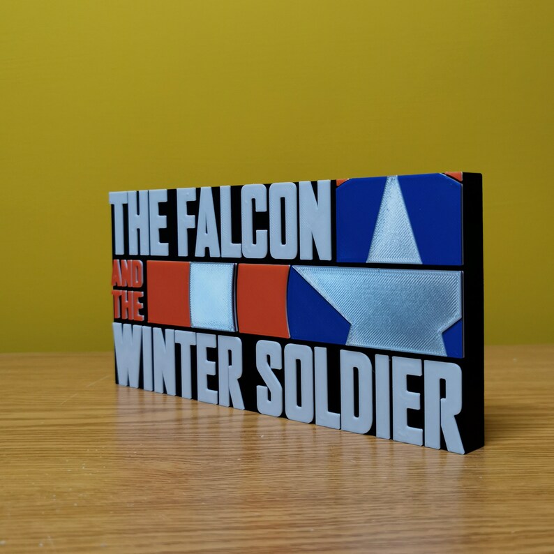 May include: A black and white 3D printed sign with the text "The Falcon and the Winter Soldier" in white letters. The sign features a red, white, and blue American flag design.