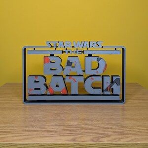 May include: A gray 3D printed sign with the text "STAR WARS THE BAD BATCH" in a black frame. The letters are in a bold, blocky font and have a red and gray speckled texture.