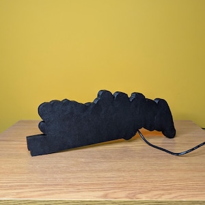 May include: A black, cloud-shaped decorative light with a warm, orange glow. The light is made of a solid material and sits on a wooden surface. A black power cord extends from the light.
