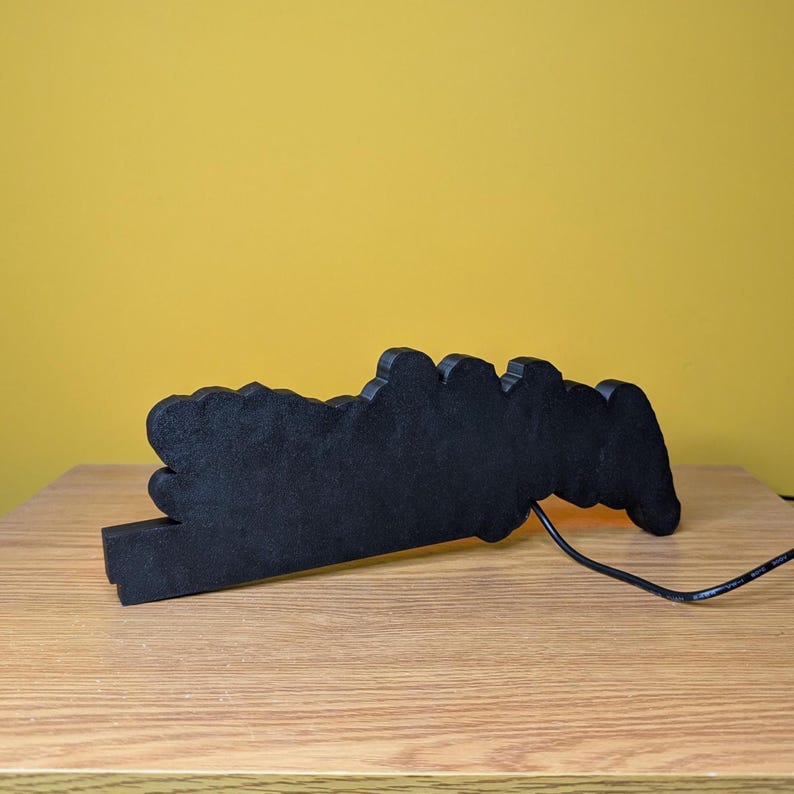 May include: A black, cloud-shaped decorative light with a warm, orange glow. The light is made of a solid material and sits on a wooden surface. A black power cord extends from the light.