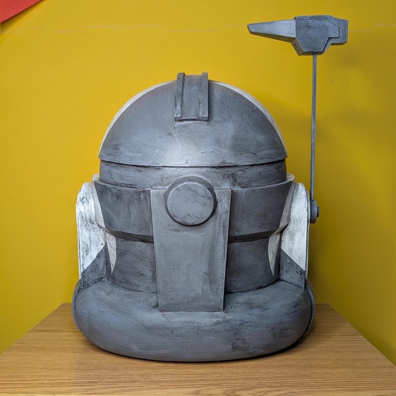 May include: A gray and white helmet with a black visor and a gray antenna. The helmet has a textured surface and is made of plastic.