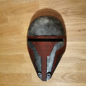 May include: A red, black, and silver helmet with a weathered finish. The helmet has a visor and a pointed chin guard.