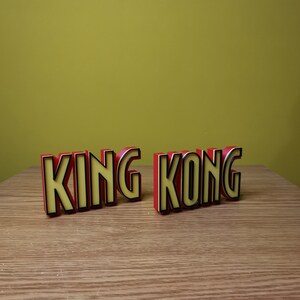 Pu&ograve; includere: Lettere a blocchi rosse e gialle che compongono la scritta "KING KONG" su una superficie di legno.