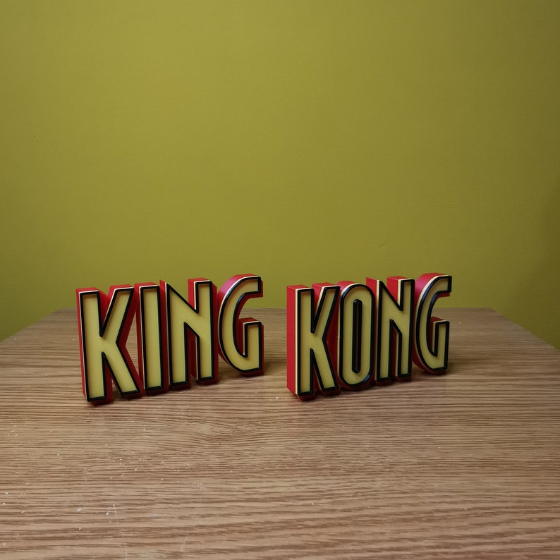 Pu&ograve; includere: Lettere a blocchi rosse e gialle che compongono la scritta "KING KONG" su una superficie di legno.