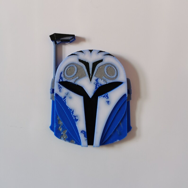 Op de afbeelding: Een blauwe en witte Mandalorian helm met een verweerd verfdesign. De helm heeft een zwart vizier en een zwarte antenne bovenop.