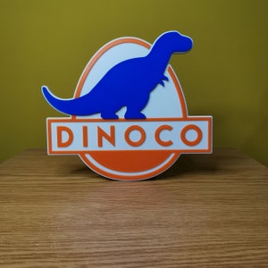 Puede incluir: Una silueta de dinosaurio azul dentro de un &oacute;valo blanco con fondo naranja y la palabra "DINOCO" en letras blancas.