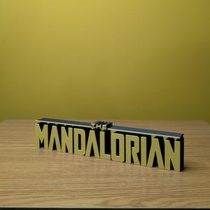 May include: A black and gold 3D printed sign that reads "THE MANDALORIAN".