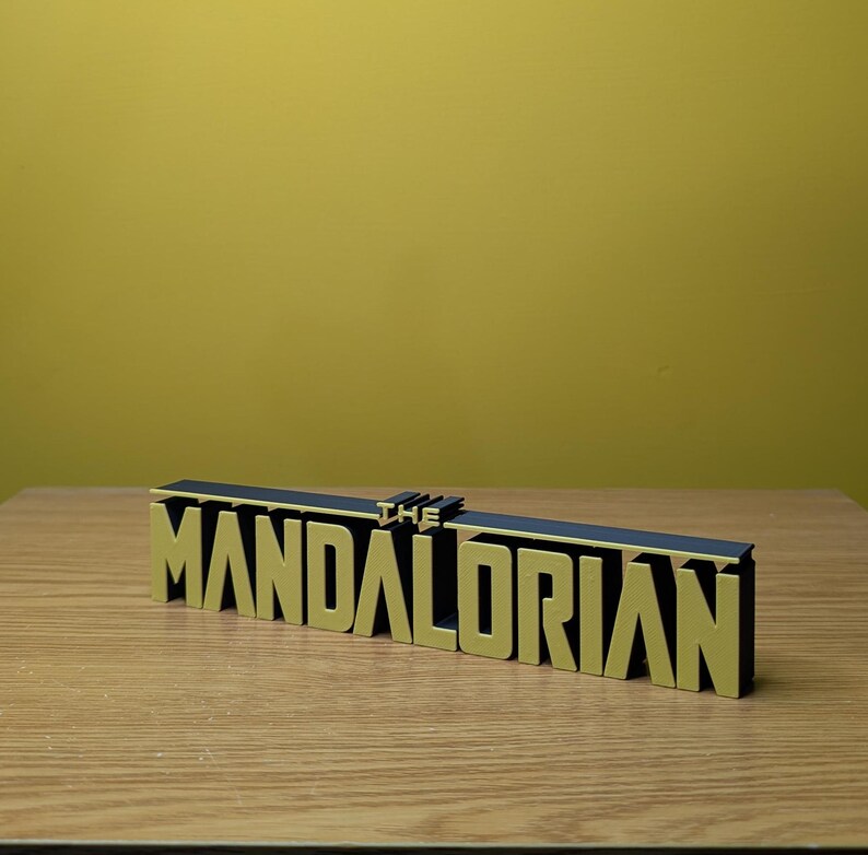 May include: A black and gold 3D printed sign that reads "THE MANDALORIAN".