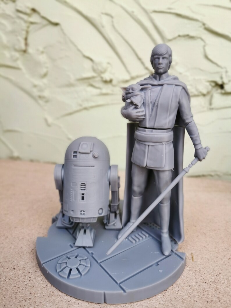 May include: A gray 3D printed figurine set featuring Luke Skywalker holding a small Yoda figure, R2-D2, and a lightsaber. The figures are standing on a circular base with a textured surface.