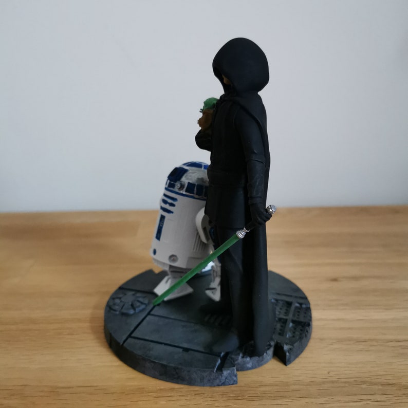 May include: A black-cloaked figure stands with a lightsaber, holding a small green figure.  A white and blue robot stands beside them on a gray base.
