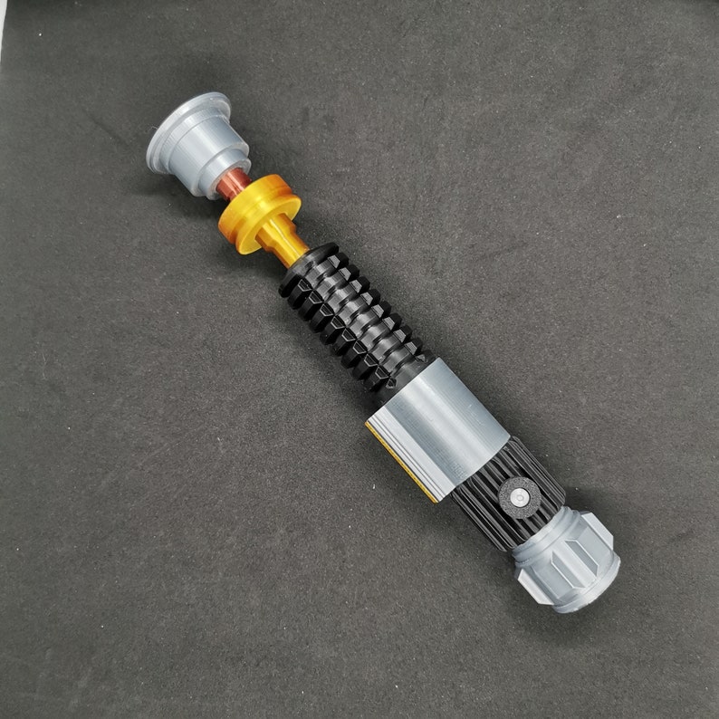 Puede incluir: Un juguete de sable de luz plateado y negro con un detalle amarillo y naranja. El sable de luz tiene un mango negro texturizado y una hoja plateada.