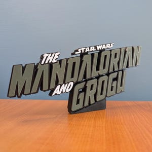 Op de afbeelding: Een 3D-geprint bord met de tekst "THE MANDALORIAN AND GROGU" en "STAR WARS" erboven. De letters zijn grijs met een textuurpatroon en zwarte contouren, gemonteerd op een zwarte basis. Het bord staat op een houten oppervlak.