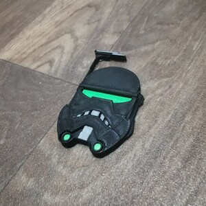 Pu&ograve; includere: Un supporto per telefono stampato in 3D di colore nero e verde a forma di casco di Stormtrooper di Star Wars.