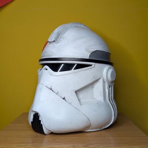 May include: A white and gray helmet with black accents, featuring a red stripe. The helmet has a weathered appearance and is set against a yellow background. The design is reminiscent of a clone trooper helmet.