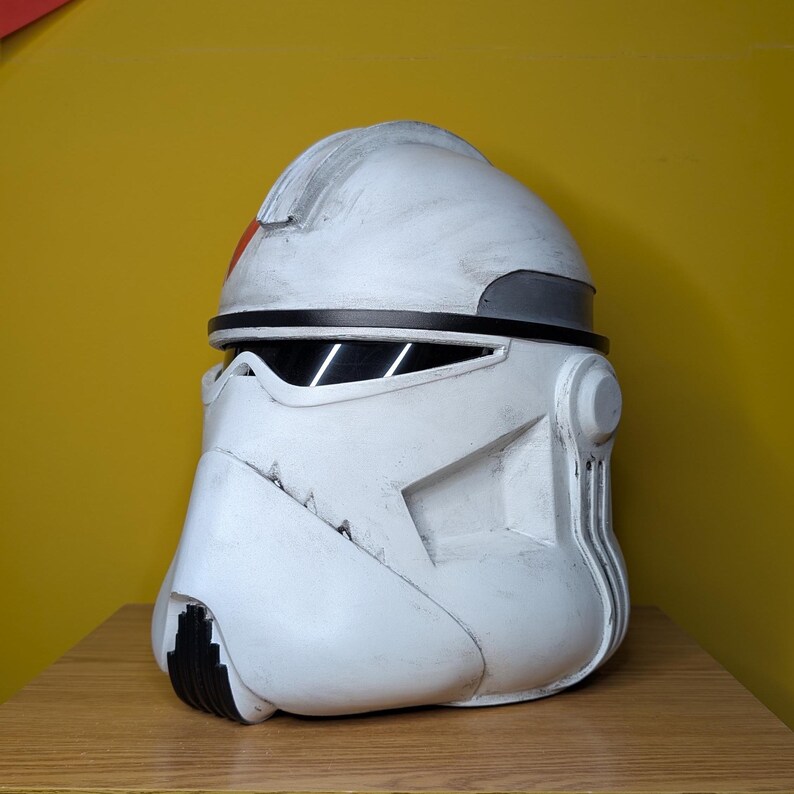 May include: A white and gray helmet with black accents, featuring a red stripe. The helmet has a weathered appearance and is set against a yellow background. The design is reminiscent of a clone trooper helmet.