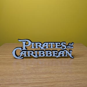 May include: A white 3D printed sign that reads "Pirates of the Caribbean" in a stylized font. The sign is on a wooden surface.