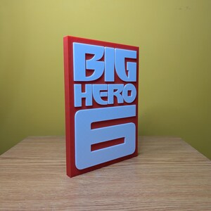 May include: A red and white 3D printed sign with the words "BIG HERO 6" in white letters.