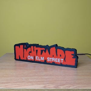 May include: A red and black sign that reads "A Nightmare on Elm Street". The sign is a 3D printed design with a black border and a red interior. The sign is sitting on a wooden surface.