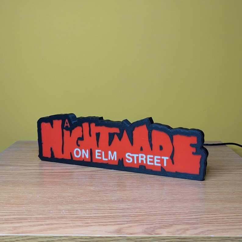 May include: A red and black sign that reads "A Nightmare on Elm Street". The sign is a 3D printed design with a black border and a red interior. The sign is sitting on a wooden surface.