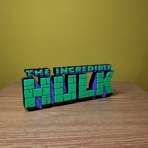 K&ouml;nnte beinhalten: Ein 3D-gedrucktes Modell des "The Incredible Hulk"-Logos. Das Logo ist aus gr&uuml;nem und violettem Kunststoff und hat eine Ziegelstruktur.