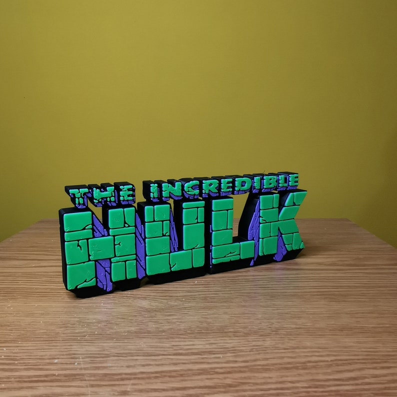 K&ouml;nnte beinhalten: Ein 3D-gedrucktes Modell des "The Incredible Hulk"-Logos. Das Logo ist aus gr&uuml;nem und violettem Kunststoff und hat eine Ziegelstruktur.