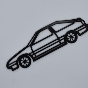 May include: Black outline of a station wagon car with visible wheels and windows. The car is angled diagonally against a white background. The wheels have a star-like design.