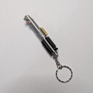 May include: A silver-colored lightsaber keychain with black and yellow accents. The keychain has a silver ring attached to the bottom. The lightsaber has a red button and a yellow rectangular detail.