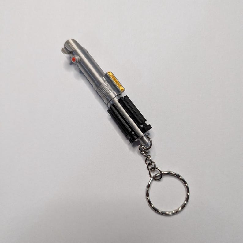 May include: A silver-colored lightsaber keychain with black and yellow accents. The keychain has a silver ring attached to the bottom. The lightsaber has a red button and a yellow rectangular detail.