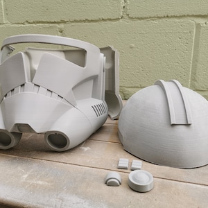 May include: A gray plastic helmet with a visor and two side vents. The helmet is in two pieces and is being assembled. There are also several smaller gray plastic pieces.