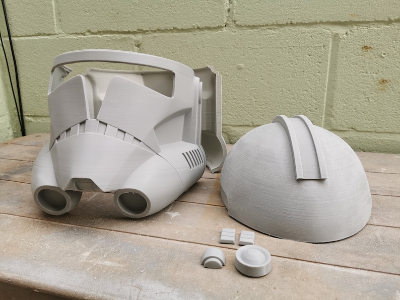 May include: A gray plastic helmet with a visor and two side vents. The helmet is in two pieces and is being assembled. There are also several smaller gray plastic pieces.