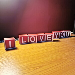 May include: Red wooden blocks spelling out the phrase "I love you" on a wooden surface.