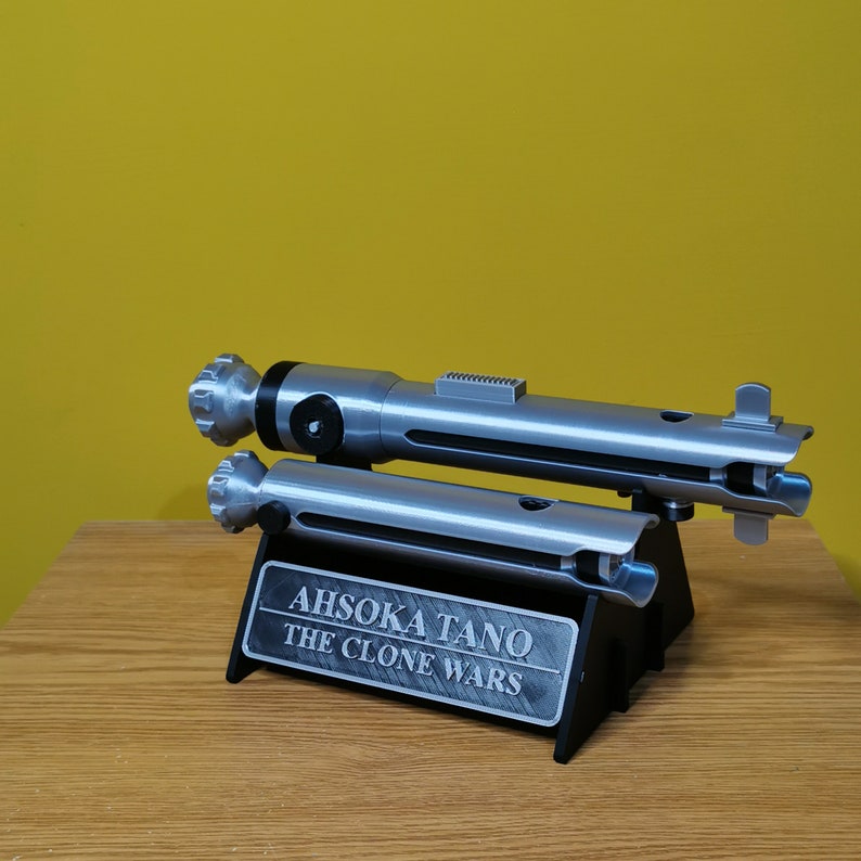 May include: Two silver lightsaber props on a black stand with a silver plaque that reads "Ahsoka Tano The Clone Wars".