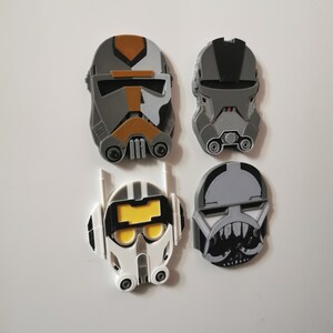 May include: Four Star Wars clone trooper helmet magnets. The helmets are gray, black, and white with yellow accents. The helmets are all different designs.