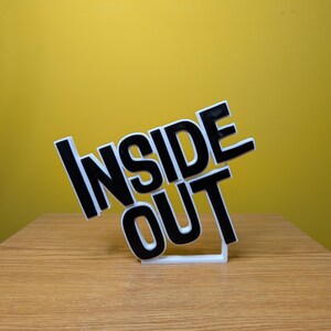 May include: A white 3D printed sign with the words "Inside Out" in black letters. The sign is tilted to the side.